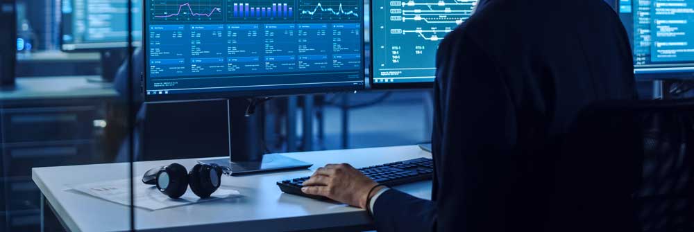 Inside a data center, see an IT professional at a computer from behind looking at two screens..