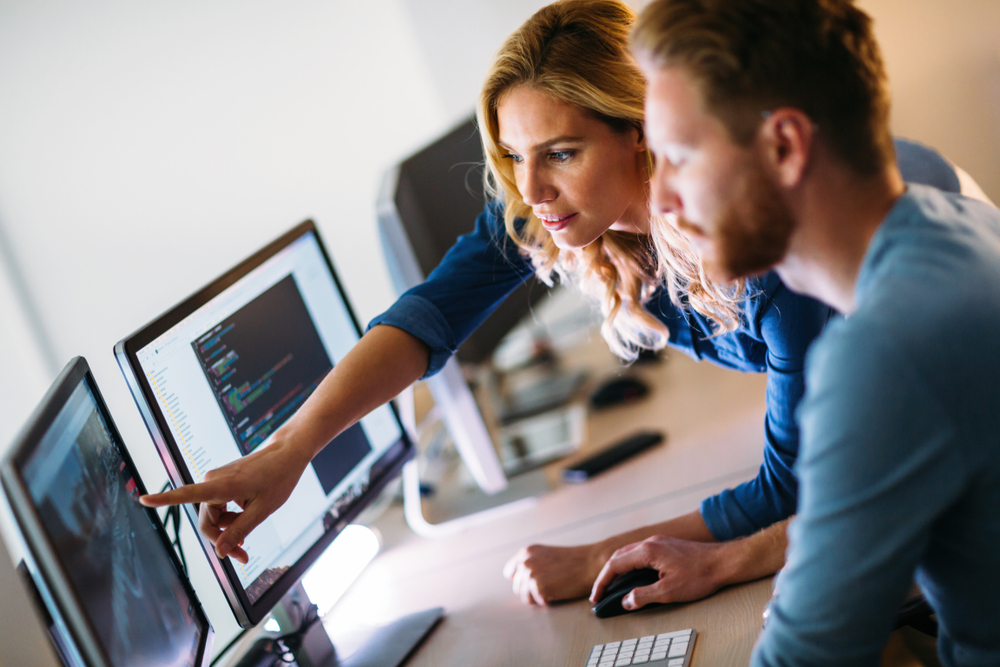 Woman pointing to a dual screen monitor to her male coworker. 