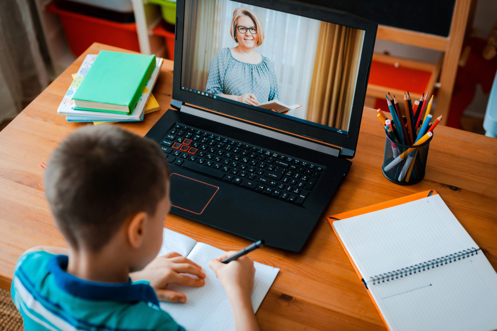 Photo from the K-12 Cybersecurity Checklist, a child is working on homework with a female teacher on screen. 