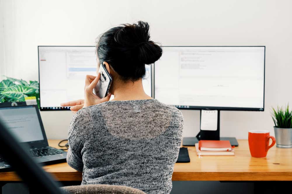 Une femme sur son téléphone portable avec deux écrans d'ordinateur en face d'elle. 