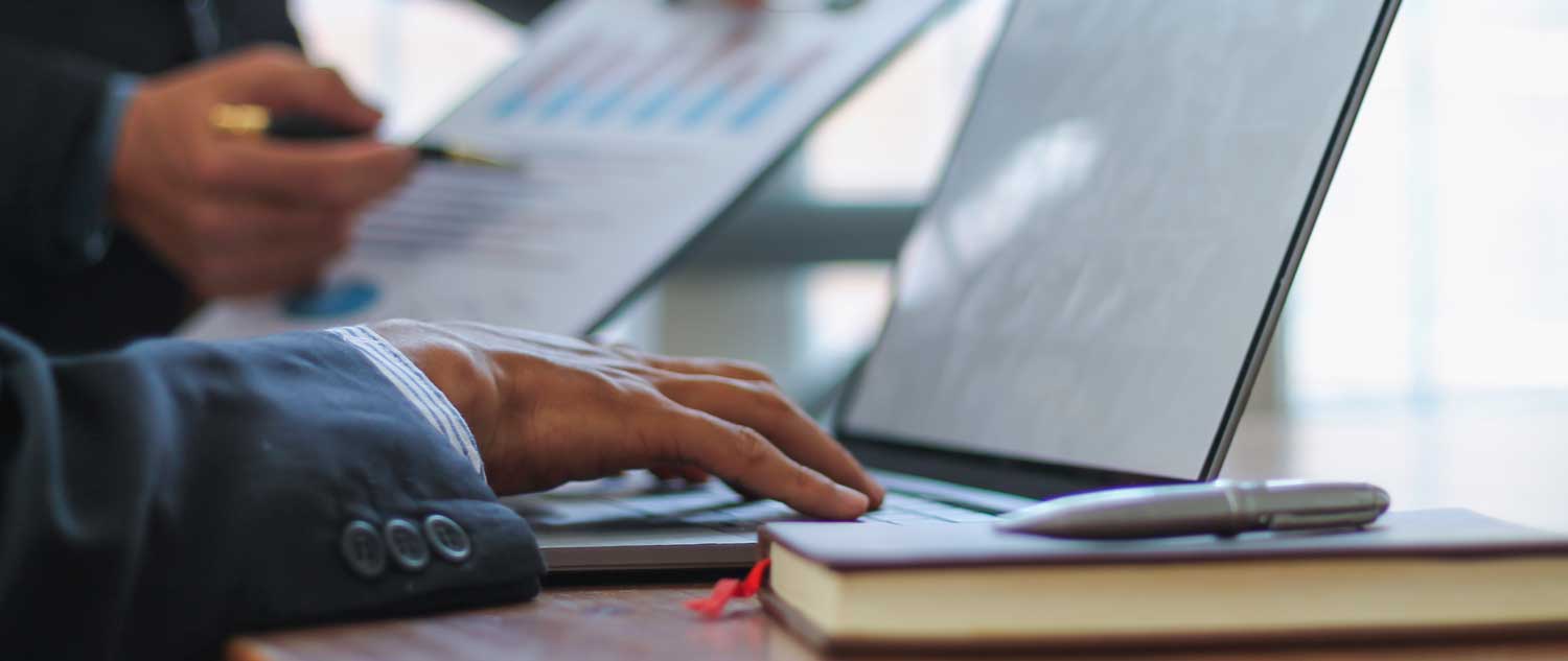 Closeup of a right hand on a laptop. A man is holding a chart on the left side of the image. 