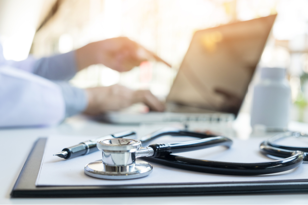 Stethoscope in the foreground with a doctor looking at his laptop in the background. 